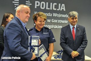 Policjantka stojąca między dwoma mężczyznami - jeden z nich trzyma ozdobna tabliczkę.