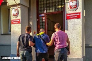 Zatrzymany mężczyzna przeprowadzany przez nieumundurowanych policjantów do budynku.