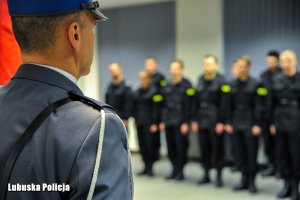 Policjant ze sztandarem policyjnym, a w tle stojacy policjanci w szeregu.