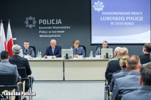 Policjanci siedzący na sali konferencyjnej, a na środku stół konferencyjny a za nim stojące osoby.