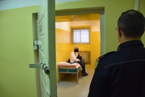 Policjant przy pomieszczeniu dla osób zatrzymanych, a w środku kobieta siedząca na łóżku.