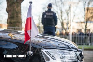 Flaga Polski na pojeździe, a w tle stojący policjant.