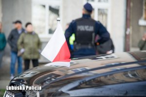 Flaga Polski na pojeździe, a w tle stojący policjant i inne osoby.