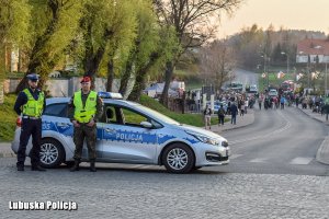 Policjant i żandarm przy radiowozie, a w tle ulica i idące chodnikiem grupy ludzi.