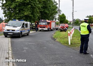 Policjant ruchu drogowego zabezpieczający drogę dojazdową na miejsce zdarzenia. W tle wóz strażacki