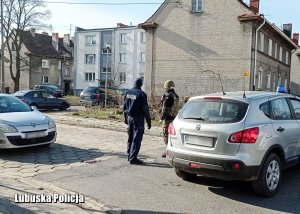 Policjant i żołnierz na tle pojazdów i budynków mieszkalnych.