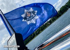 Błękitna flaga z napisem Policja na motorówce, w tle jezioro.
