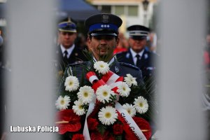 Policjant z pocztu sztandarowego z wieńcem. W tle policjant i strażak