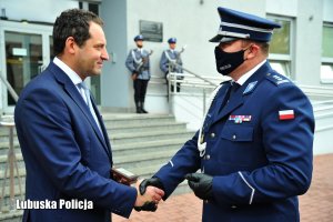 Policjant podający dłoń uczestnikowi uroczystości. W tle policjanci z posterunku honorowego