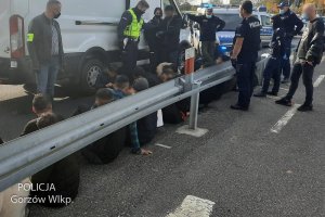 Osoby siedzące przy barierce energochłonnej - obok nich zaparkowany pojazd i policjanci.