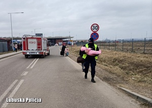 Policjant niosący dziecko, a za nimi rodzina uchodźców.