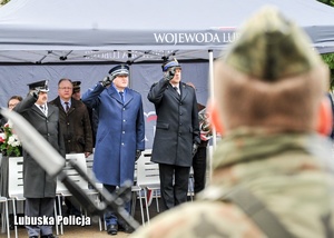 Żołnierz z karabinem, a w tle przedstawiciele służb mundurowych oddający hołd.