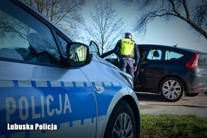 Policyjny radiowóz, a w tle policjant dokonujący kontroli drogowej.