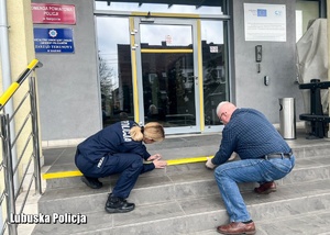 Policjantka z inną osobą nakleja taśmę na schody przed komendą.