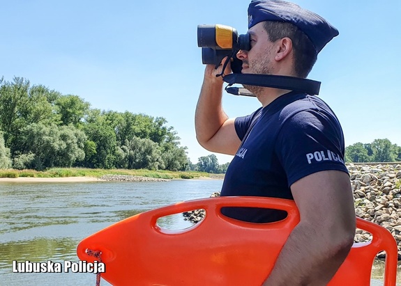 Policjant patrzy przez lornetkę, w ręce trzyma bojkę ratowniczą.