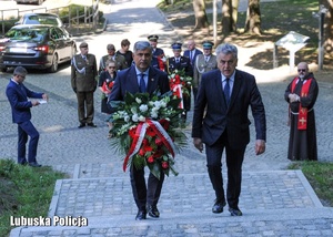 Mężczyźni idą w kierunku pomnika z wieńcem kwiatów.