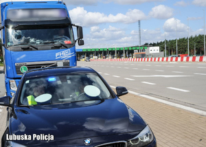 Policjanci siedzący w radiowozie nieoznakowanym, a za nimi zaparkowana ciężarówka.