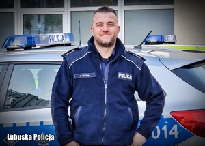 Umundurowany policjant stojący na tle radiowozu.