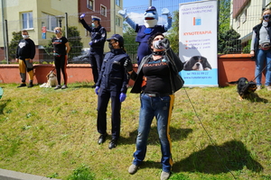 kyno terapeuta mówi przez megafon do dzieci siedzących przed oknem w szpitalu. Obok stoi policjantka, a w oddali przewodnicy psów z psami, polfinek oraz policjant