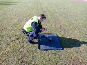 Policjant trzyma w ręku pilota od drona, który leży na ziemi przed nim