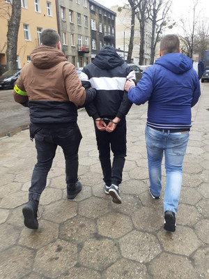 dwaj nieumundurowani policjanci prowadzą zatrzymanego skutego kajdankami założonymi na ręce trzymane z tyłu