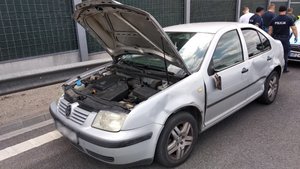 Zdjęcie przedstawia srebrny pojazd osobowy marki Volkswagen, pojazd ma uniesioną przednią klapę. Zaparkowany jest na pasie ruchu, w tle widać osłony dźwiękoszczelne trasy S8. Samochód posada liczne wgniecenia na lewym boku, ma też urwane lusterko boczne. Za nim widać grupę stojących tyłem policjantów w granatowych mundurach i ubranych po cywilnemu