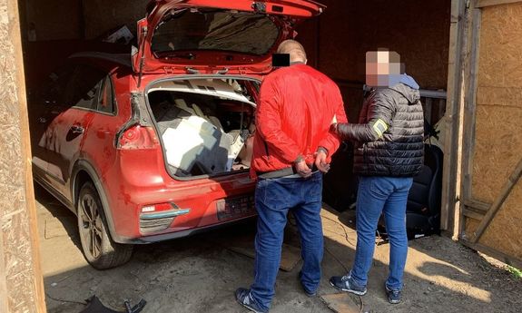 Zdjęcie przedstawia nieumundurowanego policjanta, który stoi z zatrzymanym mężczyzną przy czerwonym samochodzie zaparkowanym w garażu. Zatrzymany mężczyzna ma kajdanki założone na rekach trzymanych z tyłu.