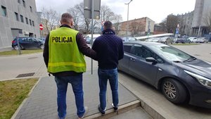 Policjant w odblaskowej kamizelce z napisem policja prowadzi zatrzymanego.