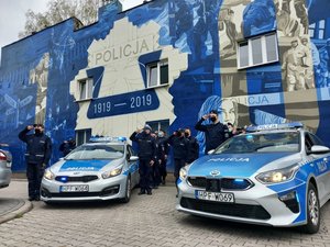 Dwa oznakowane radiowozy obok umundurowani policjanci na tle policyjnego muralu.