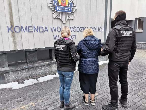 Dwoje policjantów po cywilnemu w kamizelkach wydziału kryminalnego prowadzi zatrzymaną.