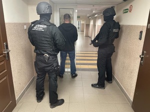 Korytarz zatrzymany stoi tyłem, za nim dwaj policjanci w mundurach, na plechach mają napis Wydział Kryminalny Komendy Wojewódzkiej Policji w Łodzi.