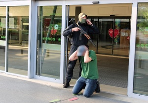Terrorysta przed wejściem do galerii trzyma broń przed nim klęczy zakładnik.