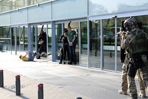 Terroryści stoją przed wejściem do galerii, mają broń zakładnicy klęczą przed nimi. Z boku idą kontrterroryści.