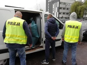 dwaj policjanci w odblaskowych kamizelkach z napisem policja stoja przed zatrzymanym białym busem razem z zatrzymanym kierowcą. Wewnatrz pojazdu widać worki