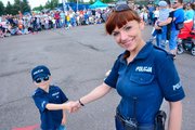 Uśmiechnięta policjantka z chłopcem, który ciągnie ją za rękę, ubrany jest w mini mundur policyjny ,w tle zgromadzeni przed pokazem goście festynu: dzieci i dorośli.