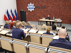 Na podeście, za stołem, w sali konferencyjnej siedzi nadinsp. dr Tomasz Michułka, Zastępca Komendanta Głównego Policji. Obok niego Pełnomocniczka Komendanta Głównego Policji ds. wdrażania Planu Równości Płci w Policji na lata 2023-2026. Na pierwszym planie uczestnicy spotkania.