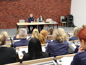 Na podeście, za stołem, w sali konferencyjnej siedzi nadinsp. dr Tomasz Michułka, Zastępca Komendanta Głównego Policji. Obok niego Pełnomocniczka Komendanta Głównego Policji ds. wdrażania Planu Równości Płci w Policji na lata 2023-2026. Na pierwszym planie uczestnicy spotkania.