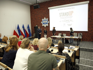Na podeście w Sali konferencyjnej Dyrektor Gabinetu Komendanta Głównego Policji wita gości - dwie kobiety, przedstawicielki Fundacji Feminoteka. Obok stoi Pełnomocniczka Komendanta Głównego Policji ds. wdrażania Planu Równości Płci w Policji na lata 2023-2026. Na pierwszym planie uczestnicy spotkania. Za osobami na podeście wyświetla się na rzutniku napis Witamy na szkoleniu. Stand Up. Sprzeciw się molestowaniu w miejscach publicznych, od L’Oréal Paris, Right To Be, Feminoteka.