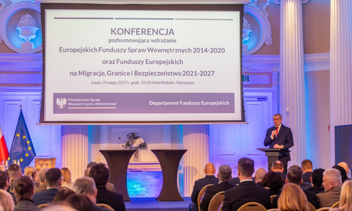 Na zdjęciu przedstawiony jest widok na salę, na której siedzą ustawieni w pięciu pięciorzędowych szeregach z prawej strony sali i trzech pięciorzędowych szeregach z lewej strony sali, odwróceni plecami, uczestnicy spotkania. Pomiędzy dwoma zgrupowaniami uczestników znajduje się przejście prowadzące na podwyższenie znajdujące się w centralnej części sali, przy tylnej jej ścianie oraz do podium z mikrofonem, które umiejscowione zostało na prawym krańcu podwyższenia - przemawia przy nim ubrany w granatowy garnitur, czerwony krawat i białą koszulę siwy mężczyzna w średnim wieku, sekretarz stanu, pan Wiesław Szczepański. Na środku podwyższenia stoją dwa dekoracyjne podia, a z jego lewej strony znajduje się flaga Unii Europejskiej oraz widoczny jest fragment polskiej flagi. Nad podwyższeniem widoczna jest duża tablica, na której wyświetlana jest nazwa konferencji wraz z umieszczonym w dolnej jej części logiem MSWiA oraz nazwą &quot;Departament Funduszy Europejskich&quot;.