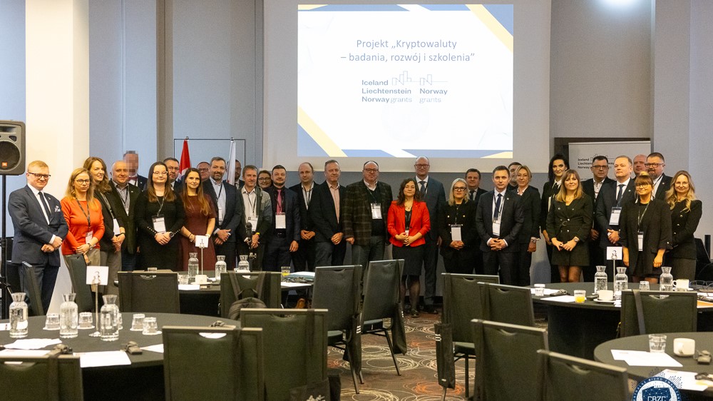 Na zdjęciu przedstawienie zostali ustawieni w poziomym rzędzie, stojący i patrzący w stronę fotografa, uczestnicy konferencji. W centralnej części fotografii, nad głowami uczestników, umieszczony został ekran, na którym wyświetlony jest slajd prezentacji PowerPoint z nazwą i logiem projektu. Na pierwszym planie zdjęcia widzimy cztery okrągłe stoliki, z pustymi krzesłami, przy których siedzieli uprzednio pozujący do zdjęcia uczestnicy konferencji. 