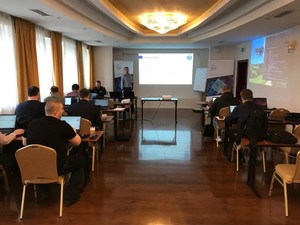 Uczestnicy szkolenia siedzą po dwie osoby przy stolikach z laptopami, prowadzący omawia prezentację.