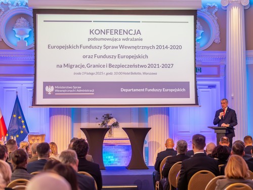 Na zdjęciu przedstawiony jest widok na salę, na której siedzą ustawieni w pięciu pięciorzędowych szeregach z prawej strony sali i trzech pięciorzędowych szeregach z lewej strony sali, odwróceni plecami, uczestnicy spotkania. Pomiędzy dwoma zgrupowaniami uczestników znajduje się przejście prowadzące na podwyższenie znajdujące się w centralnej części sali, przy tylnej jej ścianie oraz do podium z mikrofonem, które umiejscowione zostało na prawym krańcu podwyższenia - przemawia przy nim ubrany w granatowy garnitur, czerwony krawat i białą koszulę siwy mężczyzna w średnim wieku, sekretarz stanu, pan Wiesław Szczepański. Na środku podwyższenia stoją dwa dekoracyjne podia, a z jego lewej strony znajduje się flaga Unii Europejskiej oraz widoczny jest fragment polskiej flagi. Nad podwyższeniem widoczna jest duża tablica, na której wyświetlana jest nazwa konferencji wraz z umieszczonym w dolnej jej części logiem MSWiA oraz nazwą &quot;Departament Funduszy Europejskich&quot;.