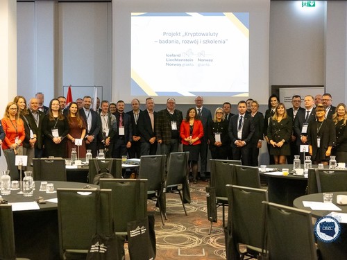 Na zdjęciu przedstawienie zostali ustawieni w poziomym rzędzie, stojący i patrzący w stronę fotografa, uczestnicy konferencji. W centralnej części fotografii, nad głowami uczestników, umieszczony został ekran, na którym wyświetlony jest slajd prezentacji PowerPoint z nazwą i logiem projektu. Na pierwszym planie zdjęcia widzimy cztery okrągłe stoliki, z pustymi krzesłami, przy których siedzieli uprzednio pozujący do zdjęcia uczestnicy konferencji.