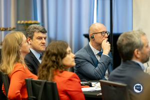 Zdjęcie przedstawia zbliżenie na siedzących przy stoliku uczestników konferencji. Znajdują się przy nim dwie widoczne z profilu kobiety ubrane z pomarańczowe żakiety oraz trzech panów w garniturach: z czego jeden widoczny jest profilem. W centralne części fotografii znajduje się obserwujący uważnie konferencję przedstawiciel partnera zagranicznego z słuchawką w uchu.