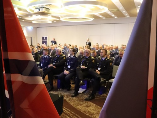 Na pierwszym planie zdjęcia, po obu jego stronach, widzimy układające się w dwa trójkąty prostokątne, tworzące prześwit w kształcie litery V ukazujący środek sali, dwie flagi. Po lewej stronie znajduje się częściowo ukazana flaga Norwegii, w drugiej - flaga o kolorze ciemnoniebieskim. 
Utworzony przez flagi prześwit ukazuje nam siedzących uczestników konferencji - policjantów z Polski i przedstawicieli norweskiego partnera.