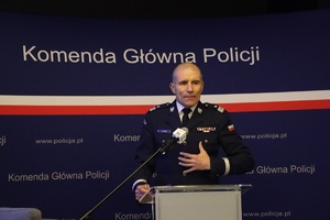 Zdjęcie przedstawia wygłaszającego przemówienie przy podium z mikrofonem Zastępcę Komendanta Głównego Policji, nadinspektora Romana Kustera w mundurze na tle ciemnoniebieskiej ścianki z białymi napisami &quot;Komenda Główna Policji&quot; umieszczonych w kilku jej miejscach oraz przechodzącym przez jej środek wąskim paskiem w barwach polskiej flagi.