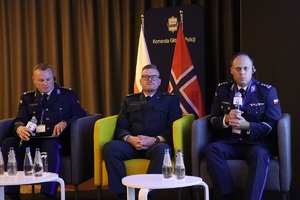 Zdjęcie przedstawia trzech członków panelu dyskusyjnego, którymi są przedstawicieli Polskiej Policji. Siedzą na trzech fotelach - boczne są niebieskie, środkowy zielony. Przed nimi znajdują się dwa małe białe stoliki, na których umieszczono butelki z wodą.
Za plecami siedzącego pośrodku uczestnika znajdują się dwie flagi - polska i norweska, za którymi z kolei znajduje się pionowa wąska ciemnoniebieska ścianka, na której umieszczono napis &quot;Komenda Główna Policji&quot;.