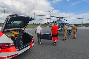 Zdjęcie przedstawia dwóch przedstawicieli transportu medycznego przenoszących skrzynię z organami do przeszczepu, obok nich znajduje się dwoje lotników. Cała czwórka zmierza do helikoptera typu Bell stojącego na lądowisku.