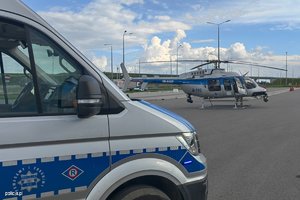 Zdjęcie przedstawia radiowóz oraz, na dalszym planie, stojący na lądowisku helikopter typu Bell.