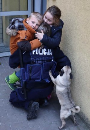 policjantka umundurowana w pozycji przyklękniętej na jedno kolano do której przytulają się dzieci  chłopiec i dziewczynka na zdjęci znajduje się również piesek